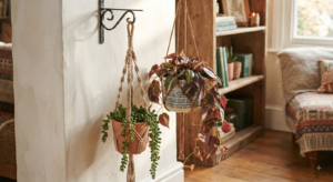 A close-up view of two artificial hanging plants in patterned and terracotta pots, suspended by macramé plant hangers from a decorative wall bracket inside a warmly lit living room.